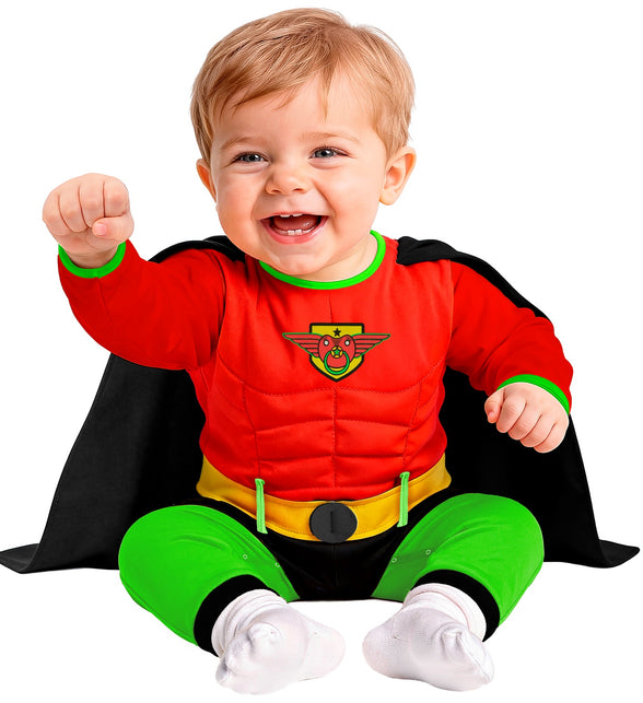 Baby in rood superheldenpak met geel schildlogo en speen, zittend met zwarte cape